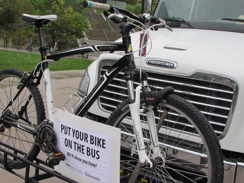 Bike rack on top of bus with sign that reads "put your bike on the bus, we'll show you how"